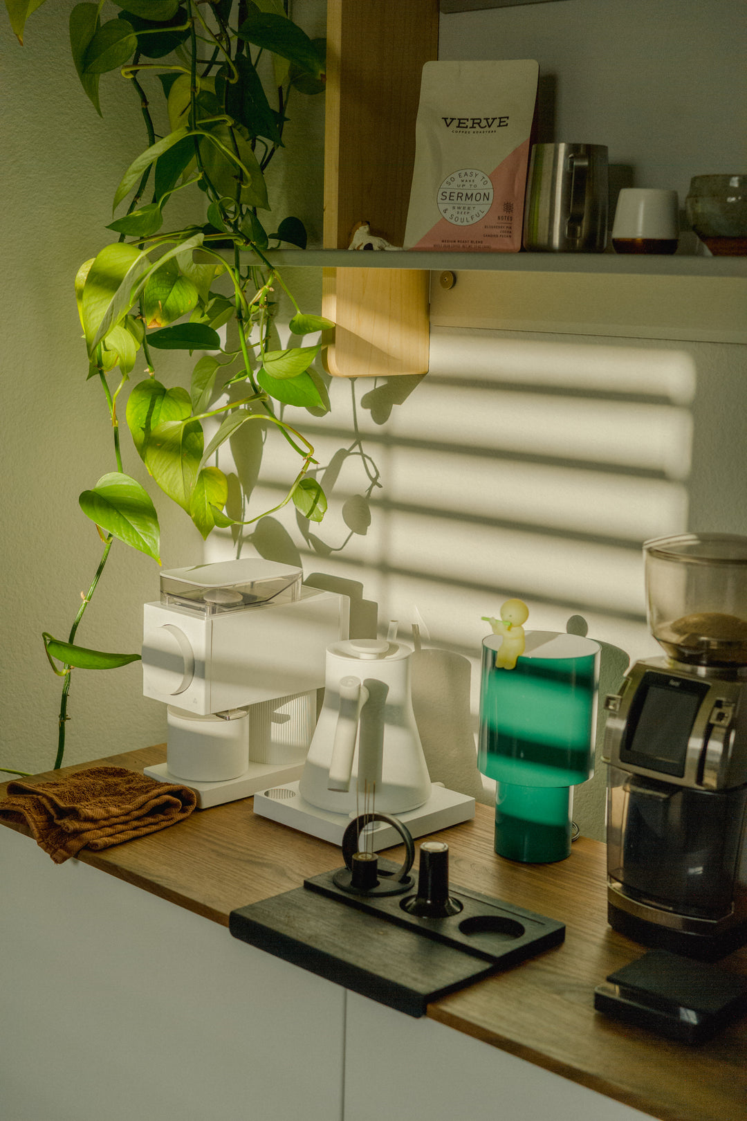 A sunlit coffee counter with grinders, a white kettle, espresso tools and wood-and-metal wall shelving