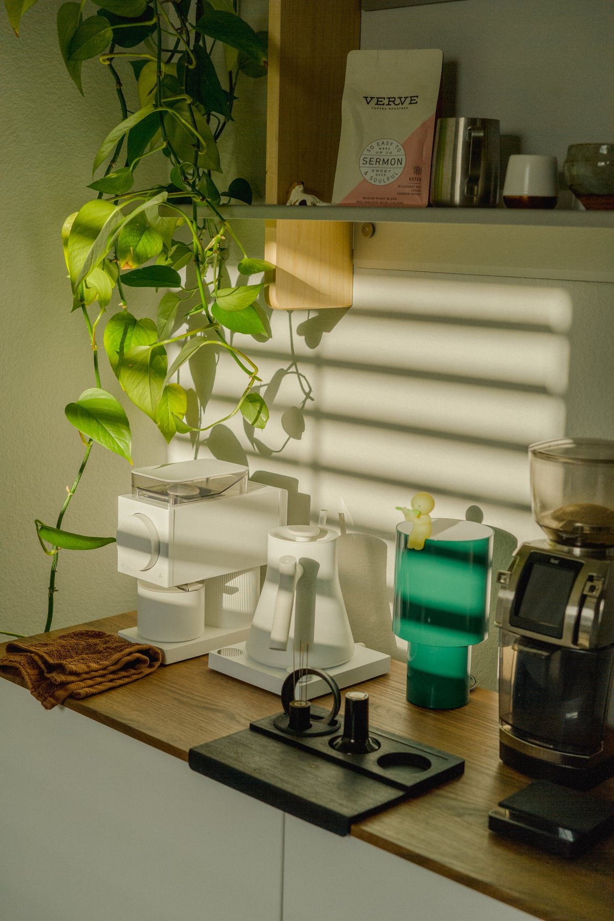A sunlit coffee counter with grinders, a white kettle, espresso tools and wood-and-metal wall shelving